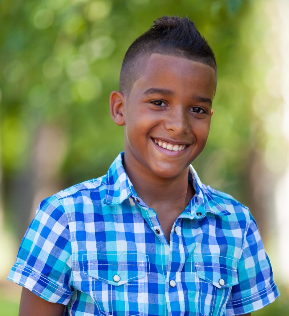Smiling boy wearing a blue and white plaid shirt stands outdoors with a blurred green background.