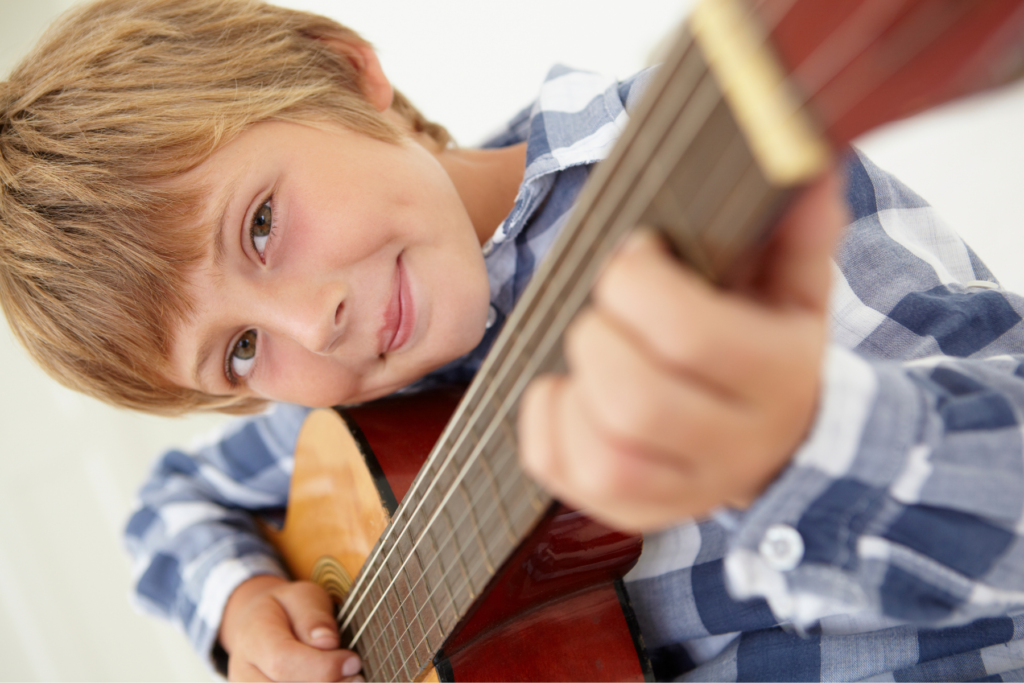Young boy in a plaid shirt playing an acoustic guitar, looking at the camera and smiling.