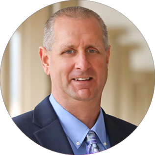 A middle-aged man with short light hair in a dark suit jacket, blue shirt, and patterned tie, standing indoors with blurred background.