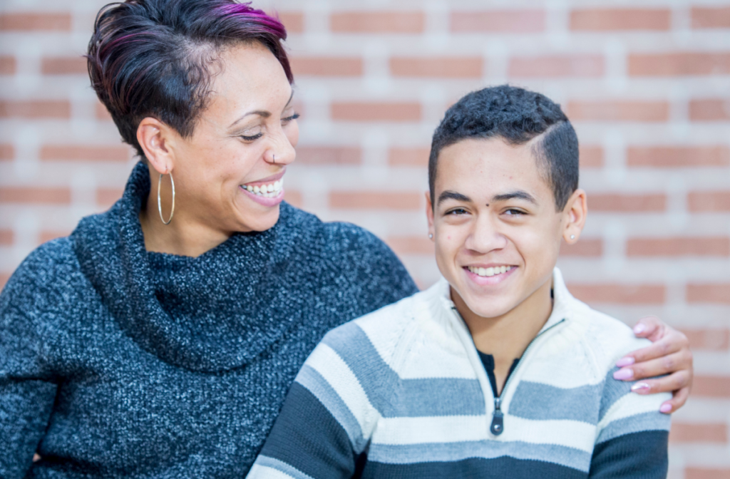 A woman and a teenage boy sit together in front of a brick wall, smiling as she puts her arm around his shoulder.