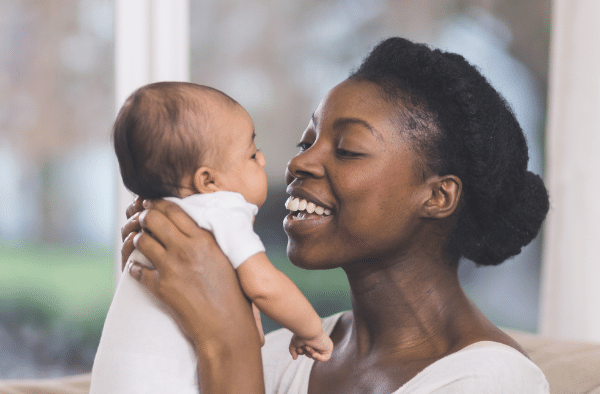 A woman holds a baby close, smiling at the child inside a bright room with a window in the background.