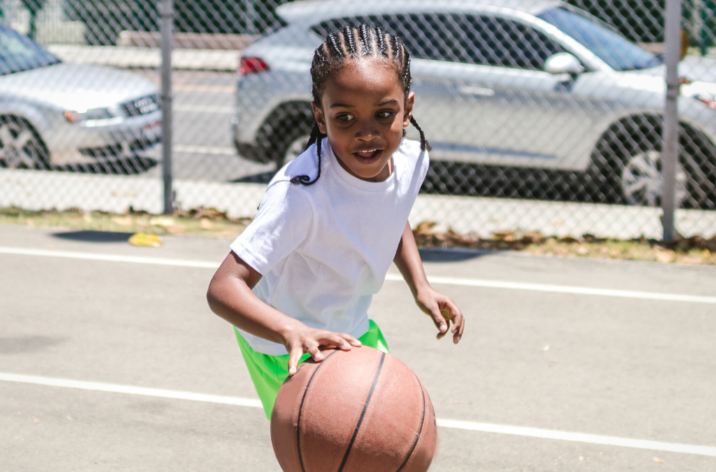 A child wearing a white shirt and green shorts dribbles a basketball on an outdoor court with cars and a chain-link fence in the background.