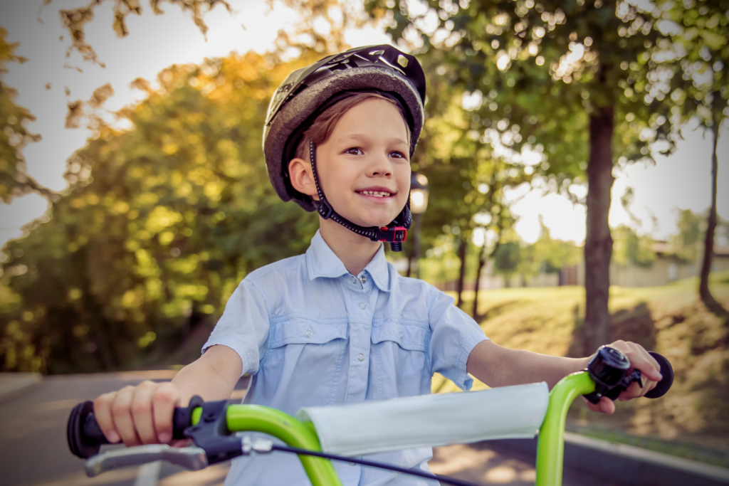 A young child wearing a helmet rides a green bicycle on a sunny day, with trees and a paved path in the background.