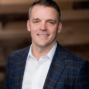 A man wearing a white shirt and a dark plaid blazer is smiling at the camera, standing in front of a blurred wooden background.