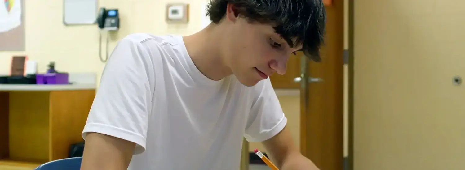 A teenage boy in a white t-shirt sits at a desk in a classroom, writing with a pencil on a piece of paper.