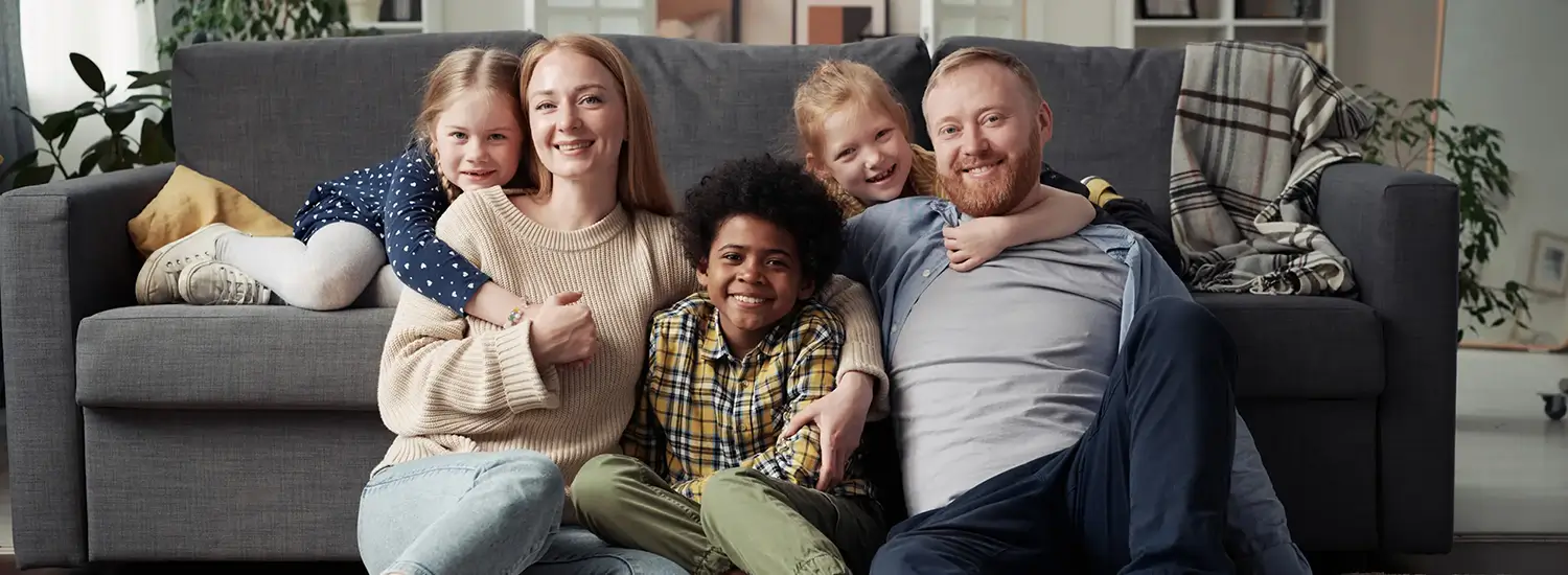 A family of five, including two adults and three children, sit together in front of a gray sofa, smiling at the camera in a cozy living room.
