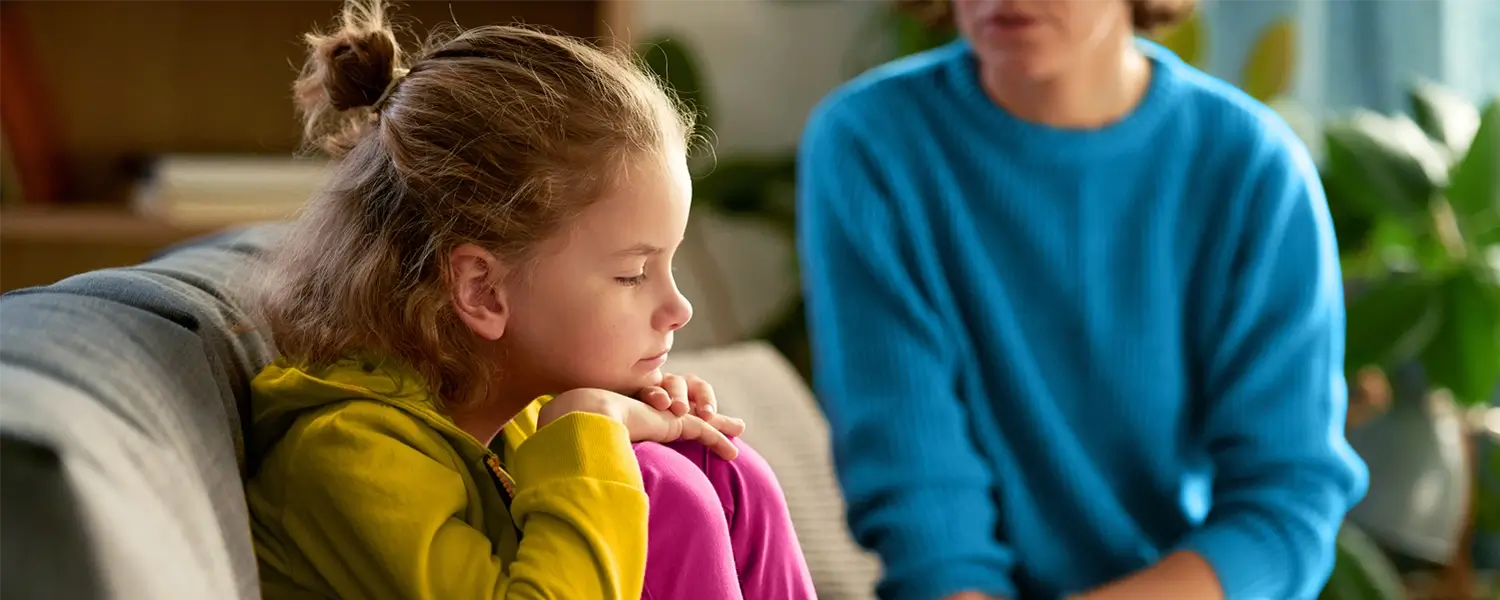 A young girl sits on a couch with her knees up, looking down thoughtfully, while an adult next to her appears to be talking.