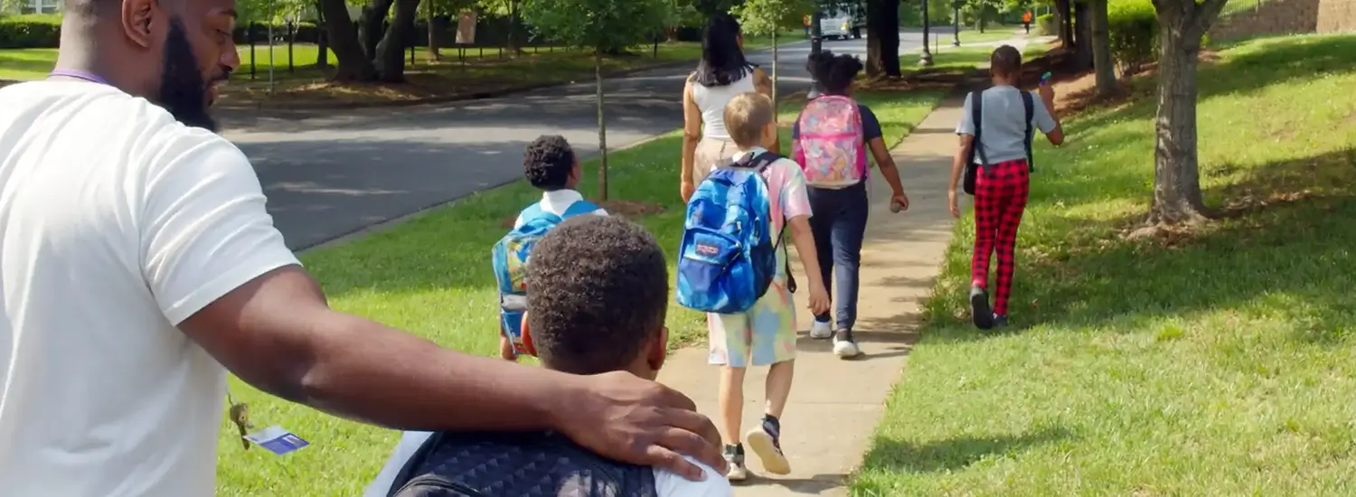 A group of children with backpacks walk on a sidewalk to school, accompanied by two adults. Trees and grass line the street.