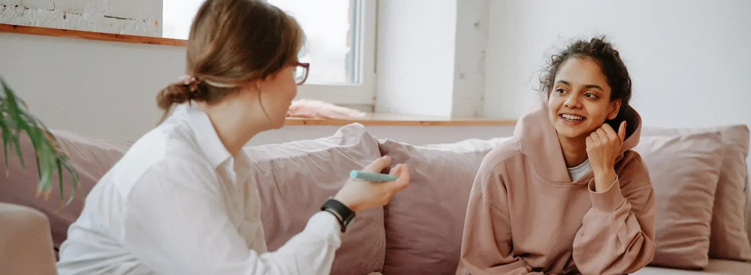 Two women sit on a couch having a conversation; one holds a pen and notebook while the other smiles and listens attentively.