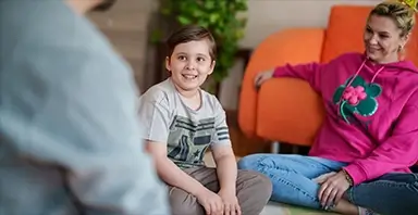 A boy sits on the floor smiling at an adult, while a woman in a pink hoodie sits nearby, both appearing relaxed in a living room setting.
