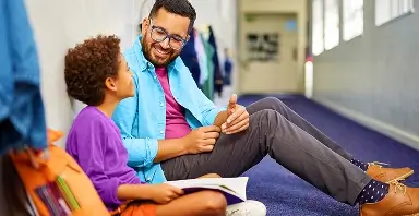 An adult and a child sit on the floor in a hallway, talking and smiling while the child holds an open book.