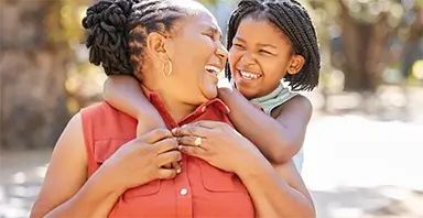 An older woman smiles while a young girl hugs her from behind and laughs; both appear happy and are outdoors in natural light.