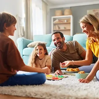 A family of four sits on the living room floor, smiling and playing a board game together.