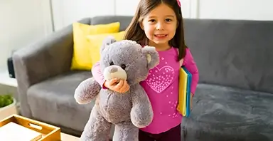 Young girl standing indoors, holding a gray teddy bear in one hand and colorful books in the other, with a gray sofa and yellow pillow in the background.