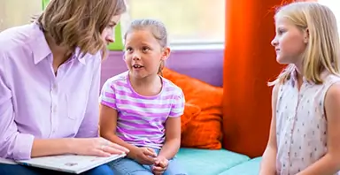 An adult woman sits with two young girls indoors, holding a folder, as they engage in conversation.
