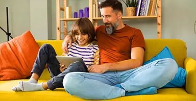 An adult and a child sit on a yellow couch, looking at a tablet together and smiling. Shelves with books and decorations are in the background.