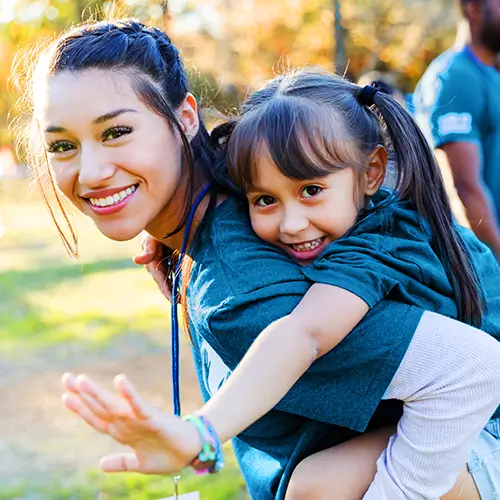 A woman gives a piggyback ride to a smiling young girl outdoors; both are wearing blue T-shirts and appear happy.