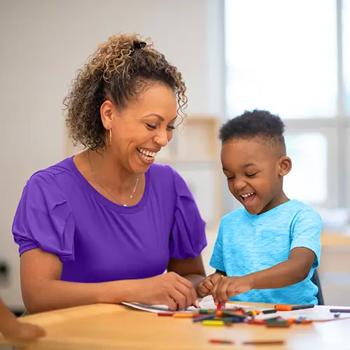 An adult and a child sit at a table, smiling and drawing with crayons together in a bright, indoor setting.
