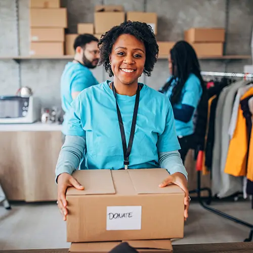 A woman wearing a blue shirt and lanyard holds a cardboard box labeled "donate" in a room with other people, boxes, and clothing racks.