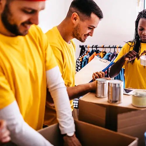 Three people in yellow shirts pack food cans into cardboard boxes, with one person holding a clipboard, in a bright room.