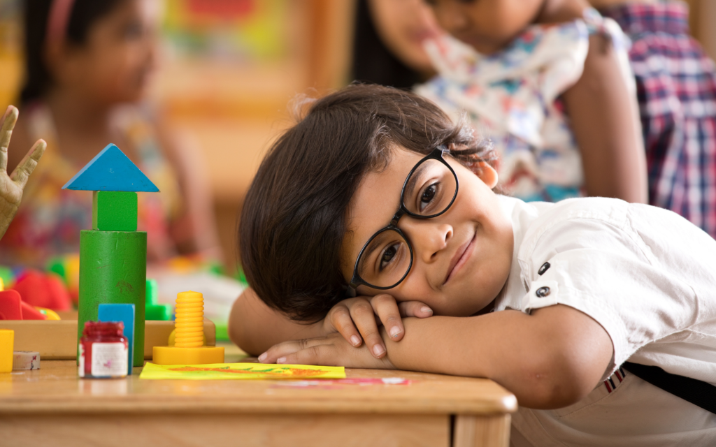 A young child wearing glasses rests their head on a desk, smiling, with colorful blocks and art supplies in the foreground and other children in the background.