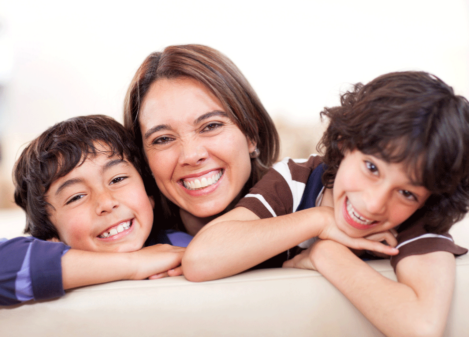 A woman smiles with two young boys, all leaning on a light-colored surface and facing the camera.
