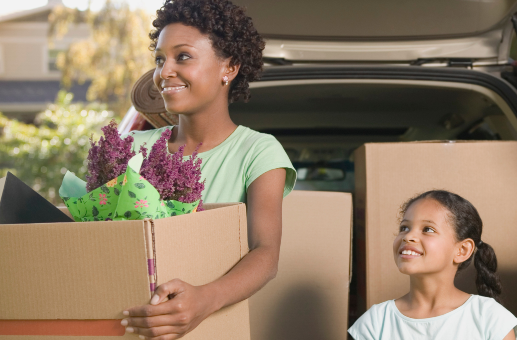 A woman carrying a box with flowers stands beside a smiling young girl in front of an open car trunk filled with moving boxes.