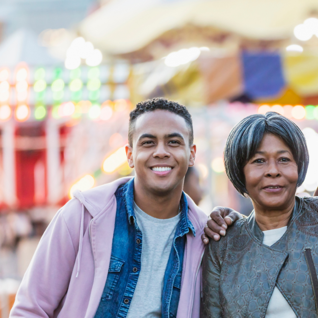 A young man and an older woman stand outdoors at a fairground, smiling at the camera with colorful lights in the background.