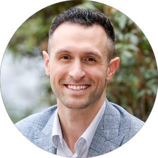 Man with short dark hair and a light gray blazer smiles at the camera, with greenery blurred in the background.
