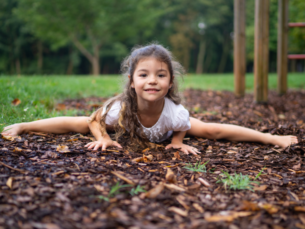 Young girl performing a front split on wood chips at a playground, smiling at the camera with green grass and trees in the background.