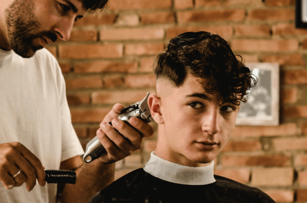 A barber uses clippers to trim the hair of a young man with a cape around his neck, against a brick wall background.