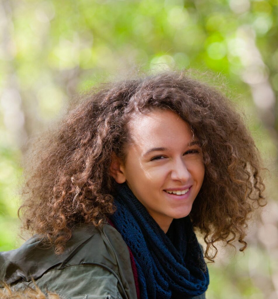 A person with curly brown hair and a dark scarf smiles outdoors with a blurred green background.