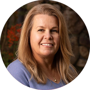 A woman with long blonde hair wearing a light blue top and a necklace, smiling, pictured outdoors with plants and rocks in the background.