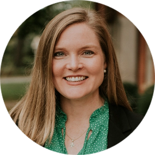 A woman with long light brown hair, wearing a green blouse and black blazer, smiles at the camera in an outdoor setting.