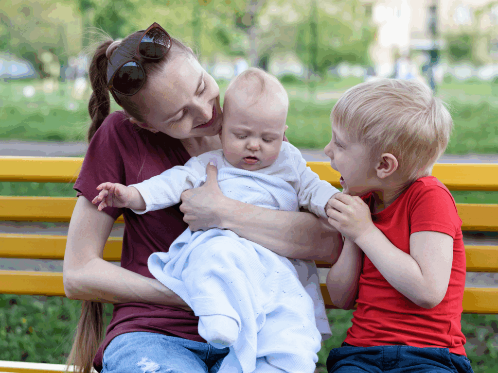 A woman sits on a yellow bench outdoors, holding a baby in a white outfit, while an older child in a red shirt sits beside them, appearing excited.