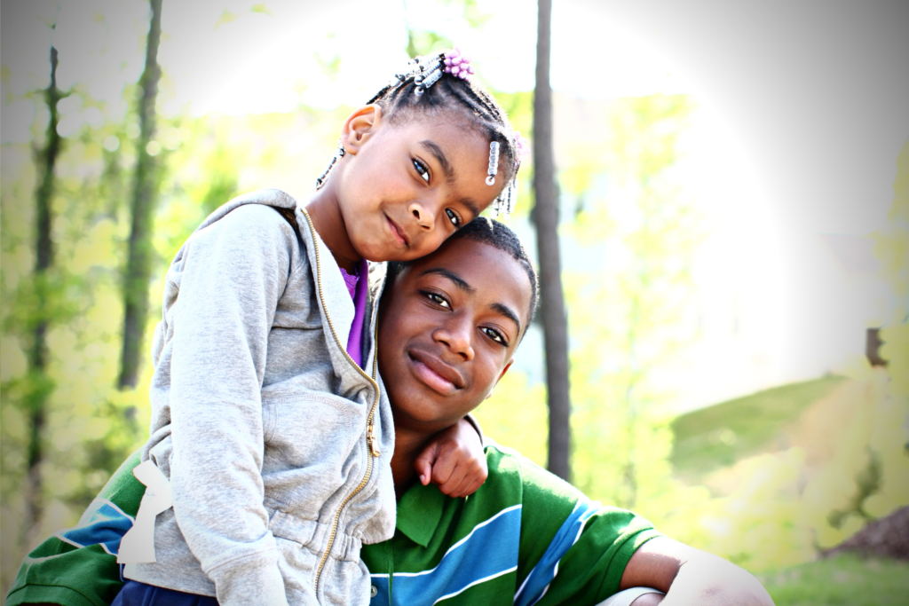 A young girl and an older boy pose together outdoors, with the girl resting her head on the boy’s shoulder and both looking at the camera.