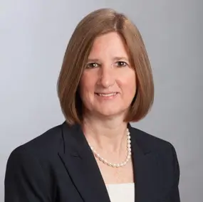 A woman with straight, shoulder-length light brown hair wearing a dark blazer, white top, and a pearl necklace, posed against a plain light background.