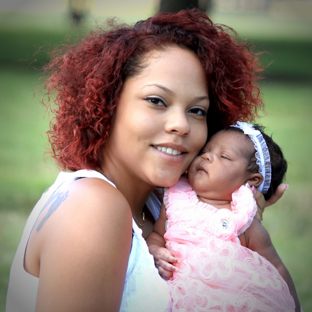 A woman with curly red hair smiles while holding a baby dressed in pink with a white headband, outdoors with a blurred green background.