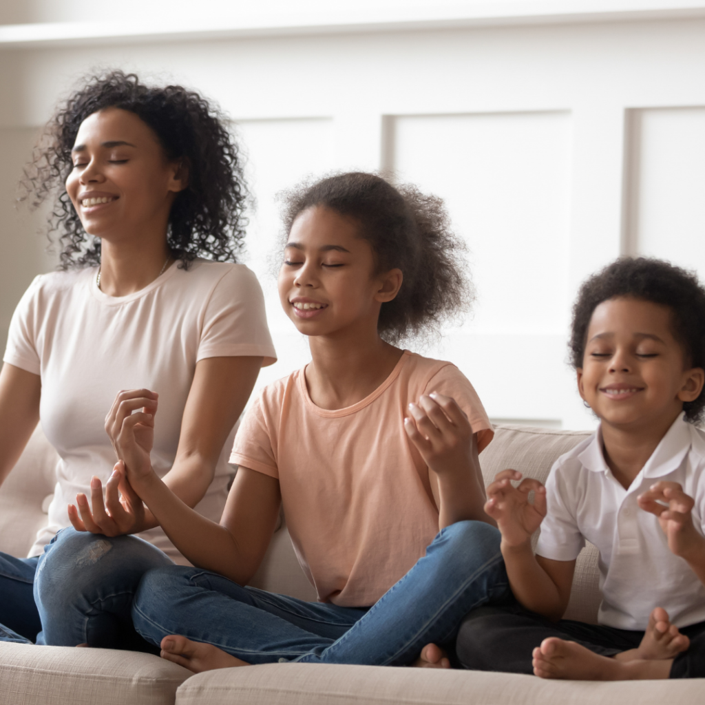 Three people sit cross-legged on a couch with eyes closed, appearing to meditate, holding their hands in a relaxed pose.