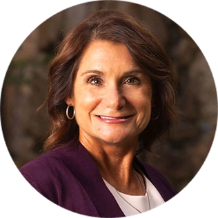 A woman with wavy brown hair, wearing a purple blazer and white top, smiles at the camera against a blurred background.