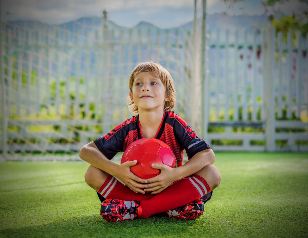 Young boy in a red and black soccer uniform sits on green turf, holding a red soccer ball, with a goal and fence in the background.