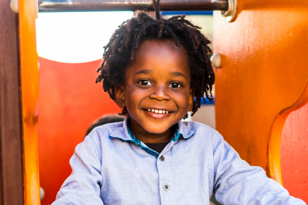 A young child with curly hair and a light blue shirt smiles while playing on orange playground equipment.