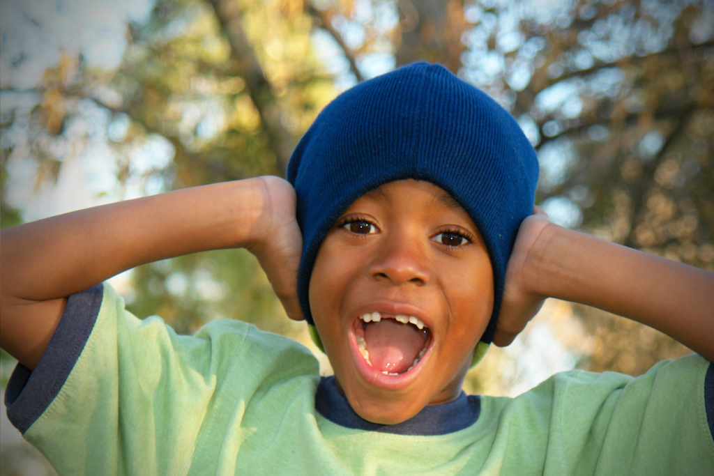 A young child wearing a blue beanie and green shirt holds their hands to their head and smiles with an open mouth. Trees and sunlight are visible in the blurry background.
