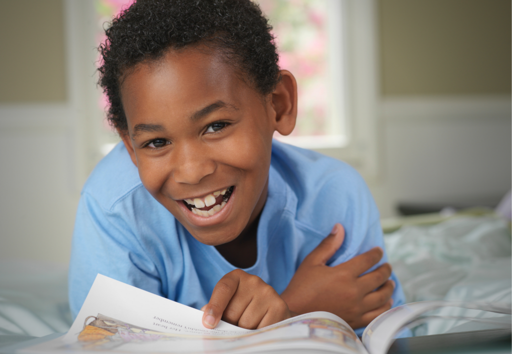 A child in a blue shirt smiles while lying down and holding an open book, with a blurred window in the background.