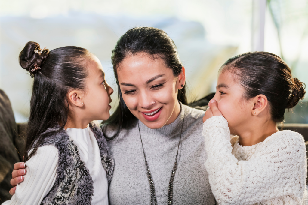 A woman sits between two young girls; one whispers into her ear while the other smiles. The trio appears to be sharing a lighthearted moment indoors.