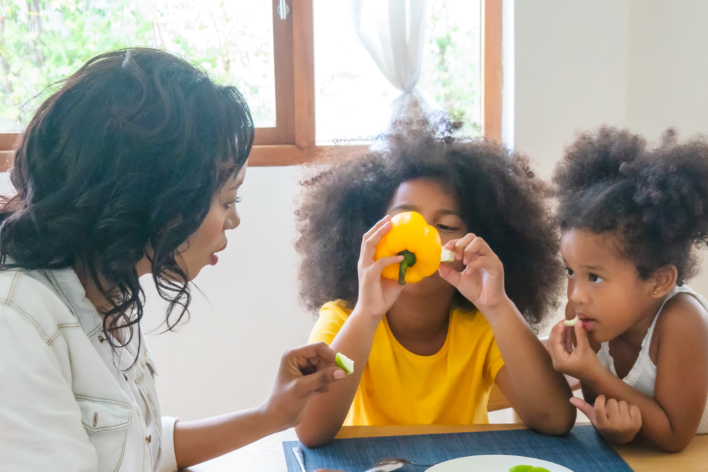 An adult and two children sit at a table. One child holds a yellow bell pepper to their face while the other watches. The adult speaks and holds a vegetable piece.
