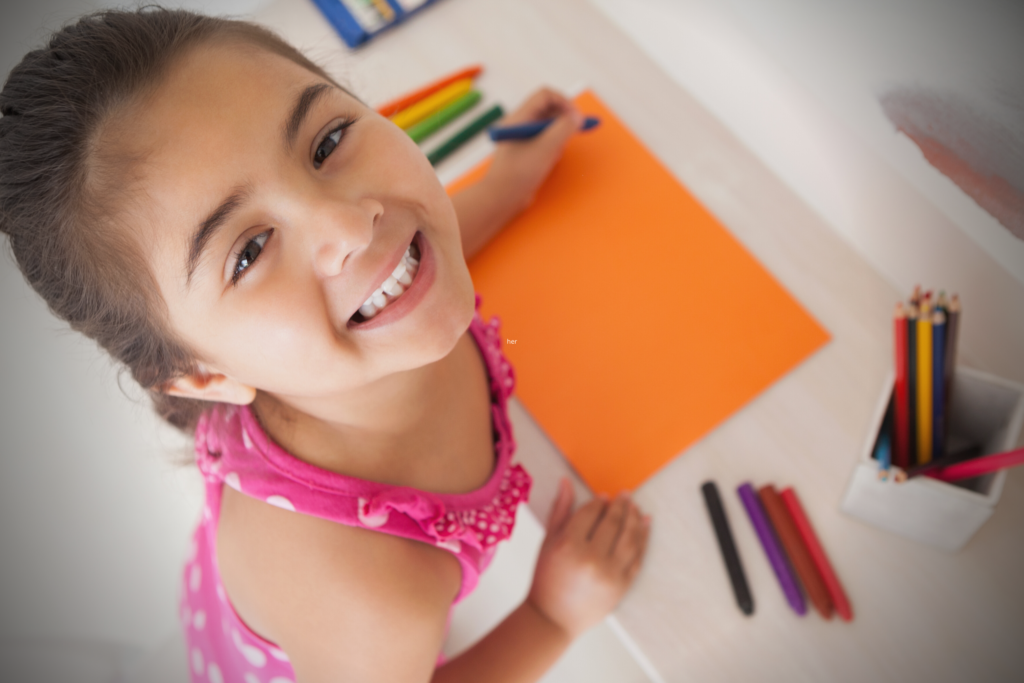 Young girl smiling up at the camera while drawing on an orange sheet of paper with colored pencils at a desk.