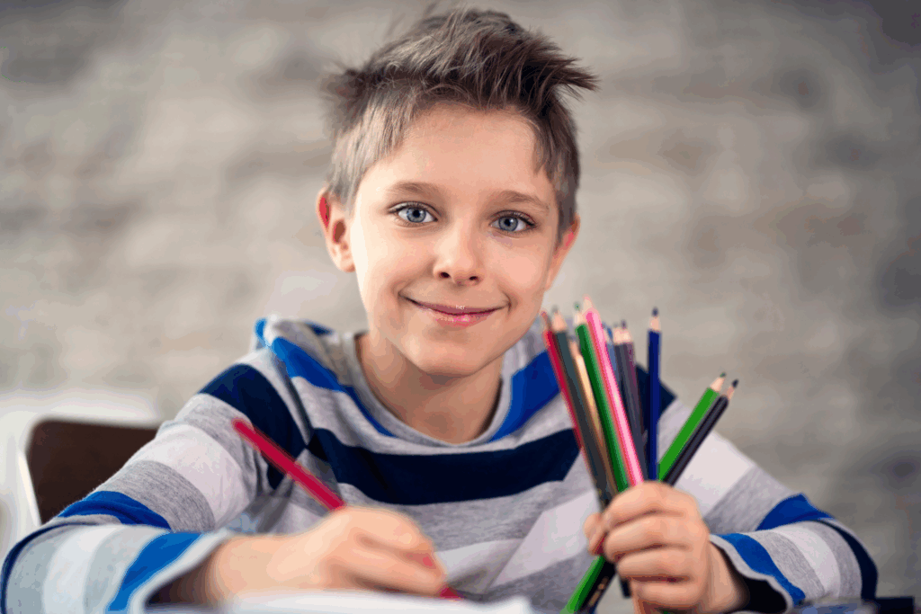 A child with short hair in a striped hoodie holds a handful of colored pencils and smiles at the camera while drawing.