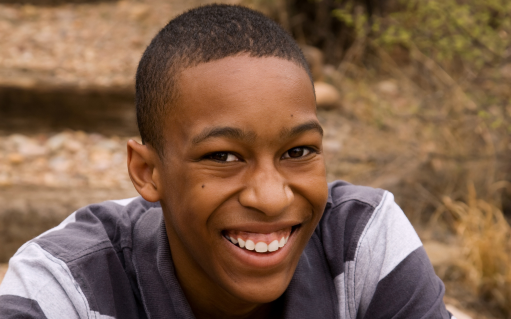 A teenage boy with short hair wearing a striped shirt smiles outdoors, with blurred steps and foliage in the background.