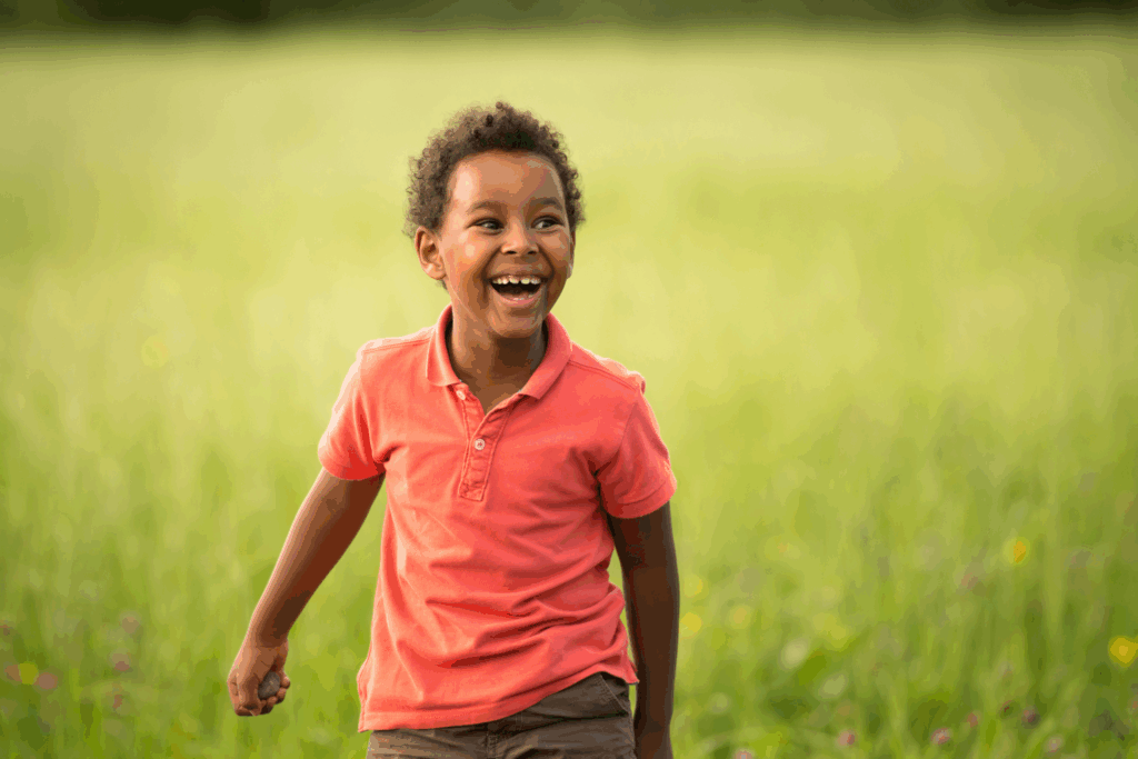A young child in a coral polo shirt smiles and stands outdoors in a grassy field with blurred green background.